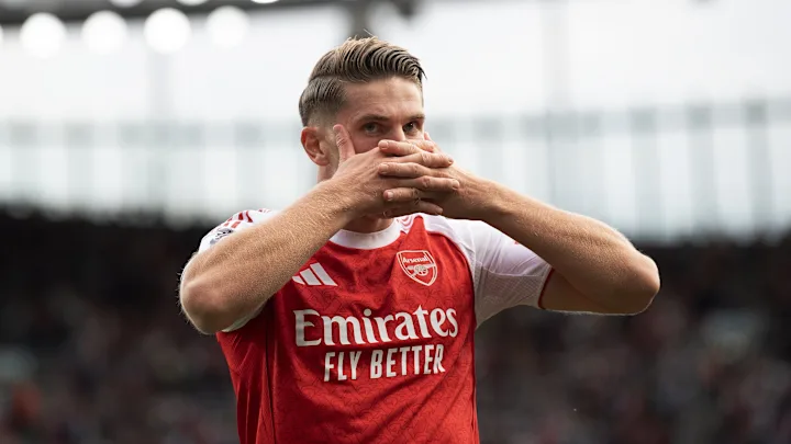 Seorang pemain Arsenal dengan seragam merah-putih merayakan gol di lapangan, tangannya direntangkan ke samping dengan ekspresi wajah gembira