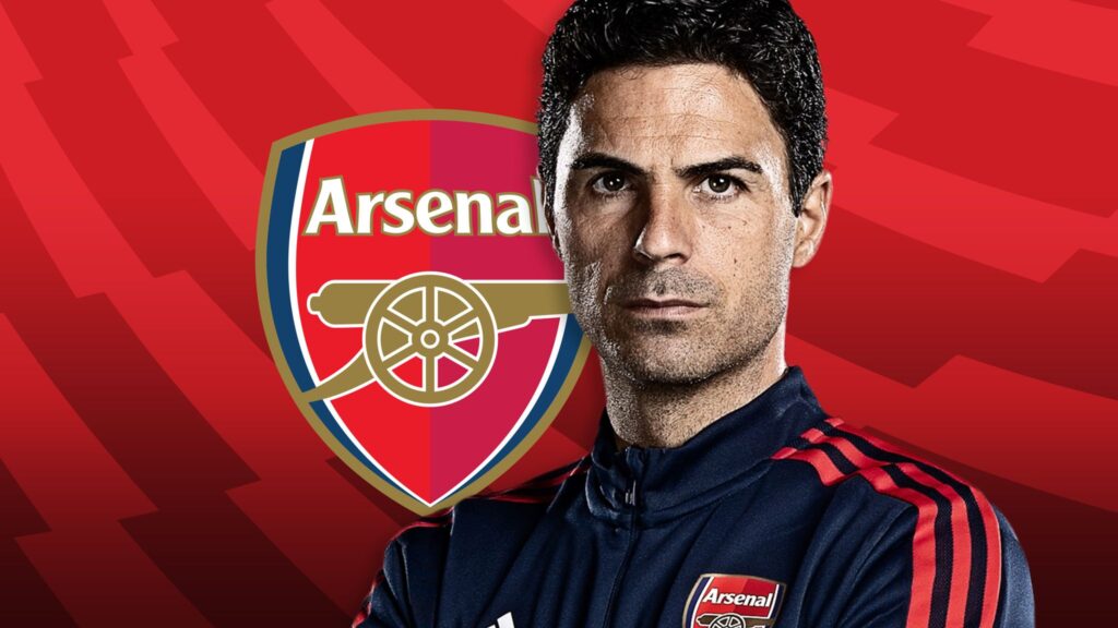 Potret Mikel Arteta, manajer Arsenal, mengenakan jaket tracksuit resmi klub berwarna merah dan biru tua, dengan latar belakang bendera atau lambang Arsenal