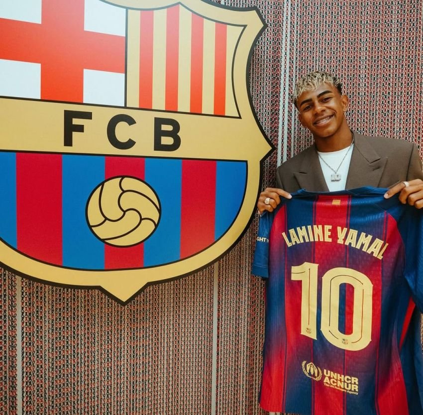 Lamine Yamal tersenyum sambil memegang jersey Barcelona bernomor punggung 10 di depan logo klub. Jersey tersebut menandakan bahwa ia adalah pewaris nomor legendaris tersebut