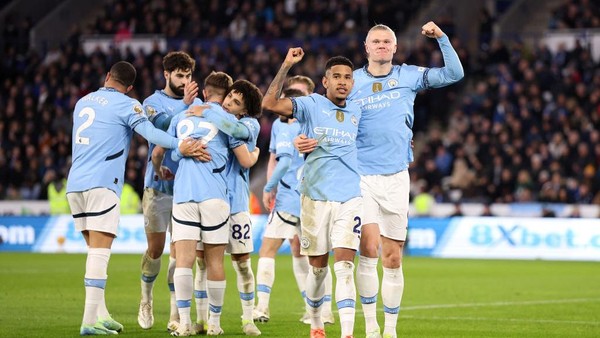 Para pemain Manchester City merayakan gol bersama, mengangkat tangan di lapangan, mengenakan jersey biru muda.