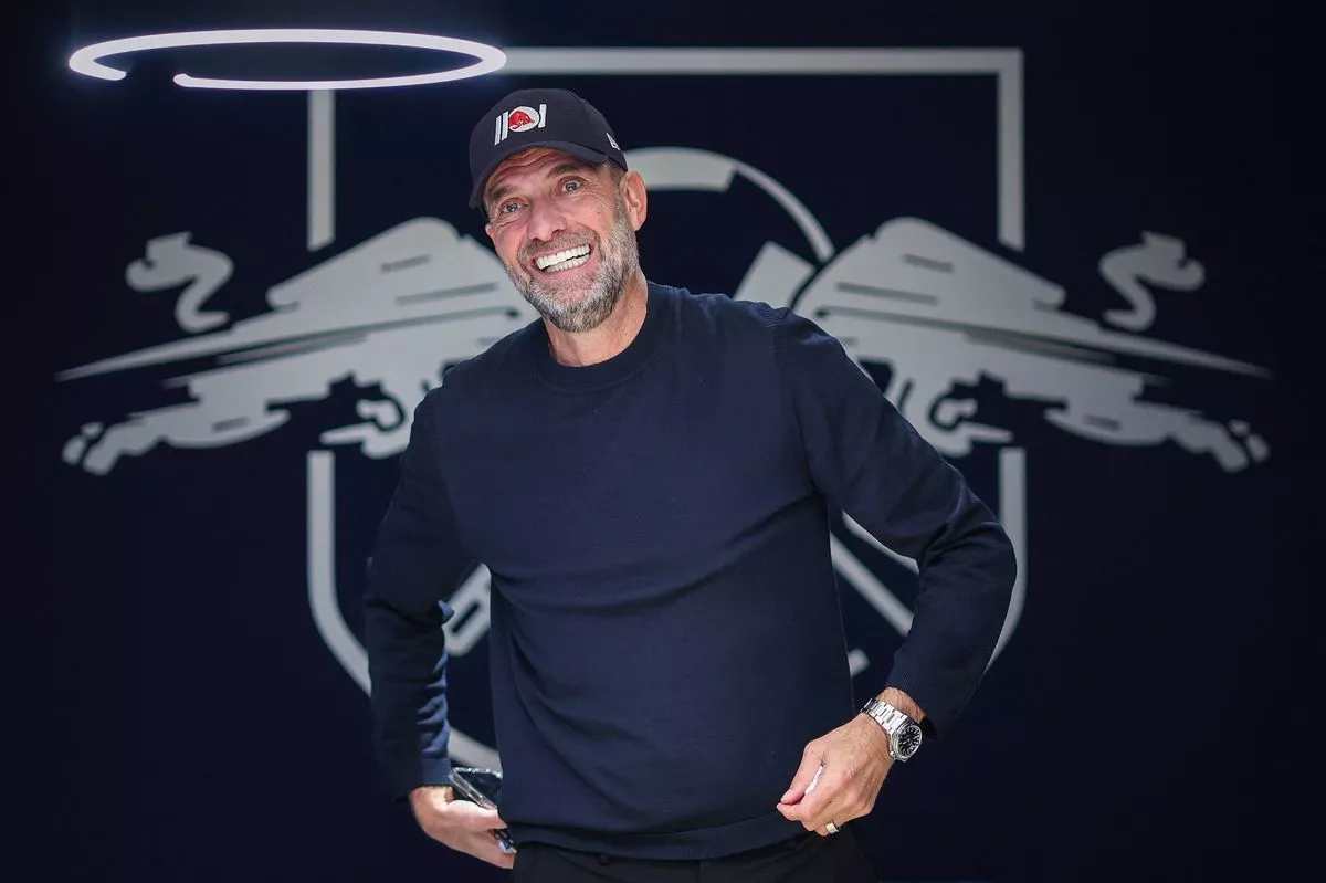 Pelatih sepak bola berdiri di depan logo klub dengan ekspresi santai