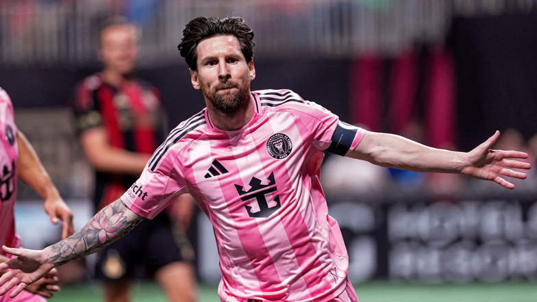 Lionel Messi mengenakan jersey pink Inter Miami sedang beraksi di tengah pertandingan dengan tangan terbuka.