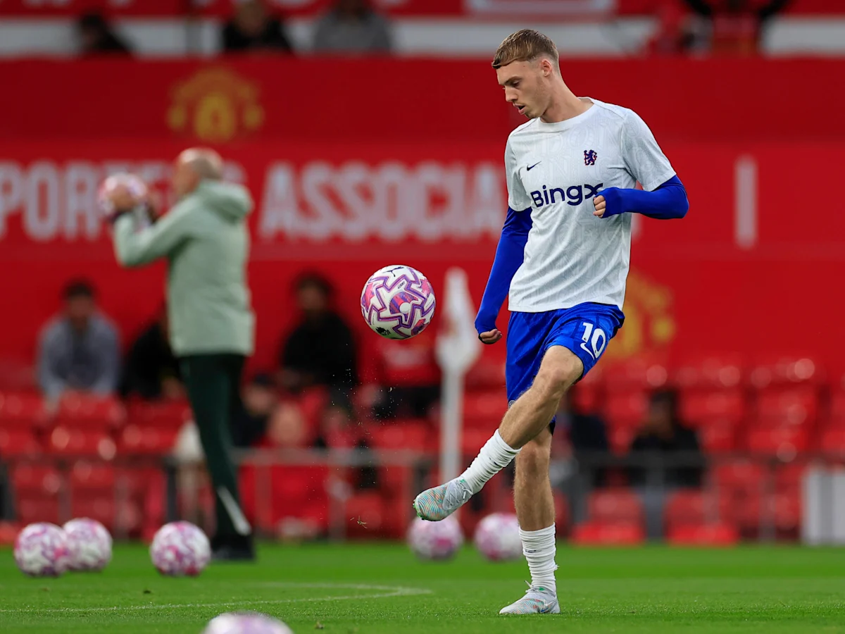 Cole Palmer, mengenakan kaus latihan putih dan celana pendek biru Chelsea, sedang menendang bola di udara di lapangan latihan dengan latar belakang kursi stadion merah.