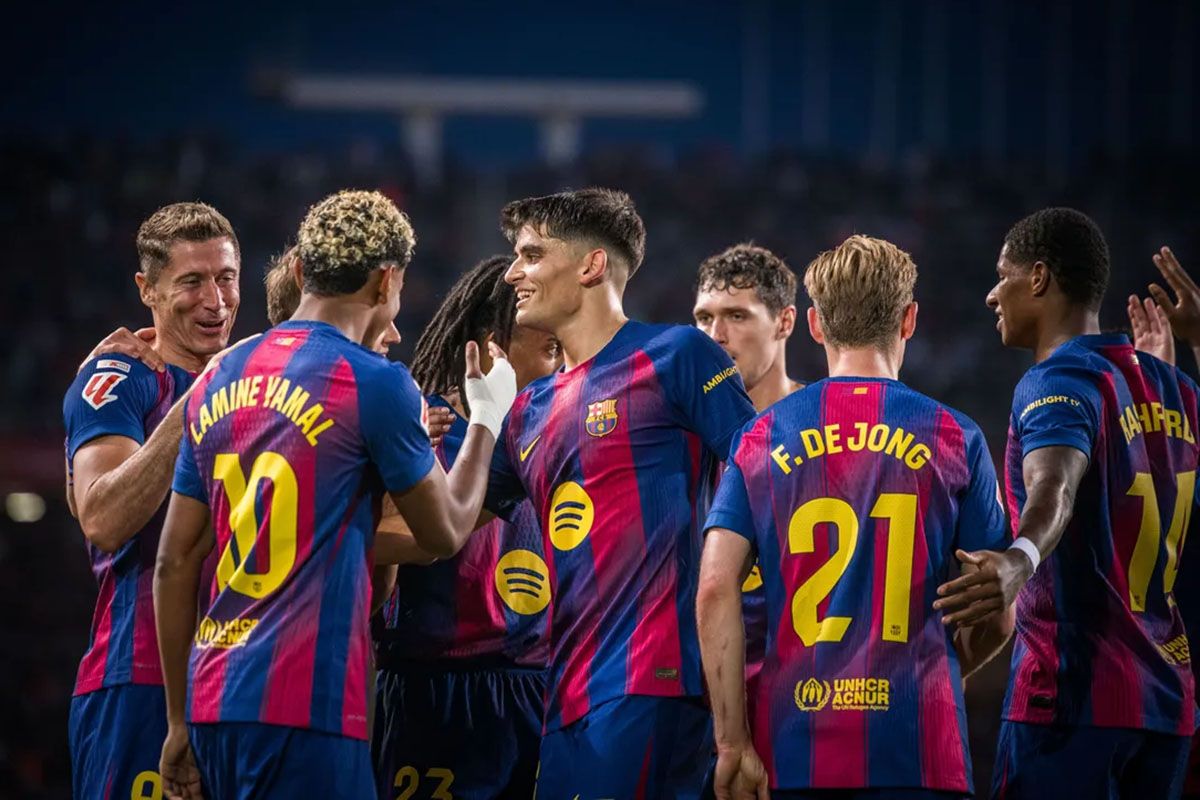 Para pemain FC Barcelona dalam balutan jersei kandang berwarna merah-biru sedang berkumpul di lapangan, dilihat dari sisi belakang, dengan fokus pada nomor punggung 21 dan rekan satu tim yang saling merangkul.