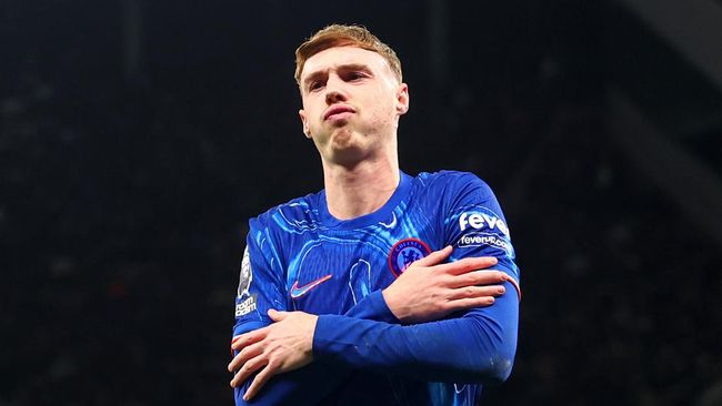 Foto setengah badan Cole Palmer dengan jersey Chelsea biru, berpose menyilangkan tangan di depan dada dengan latar belakang gelap.