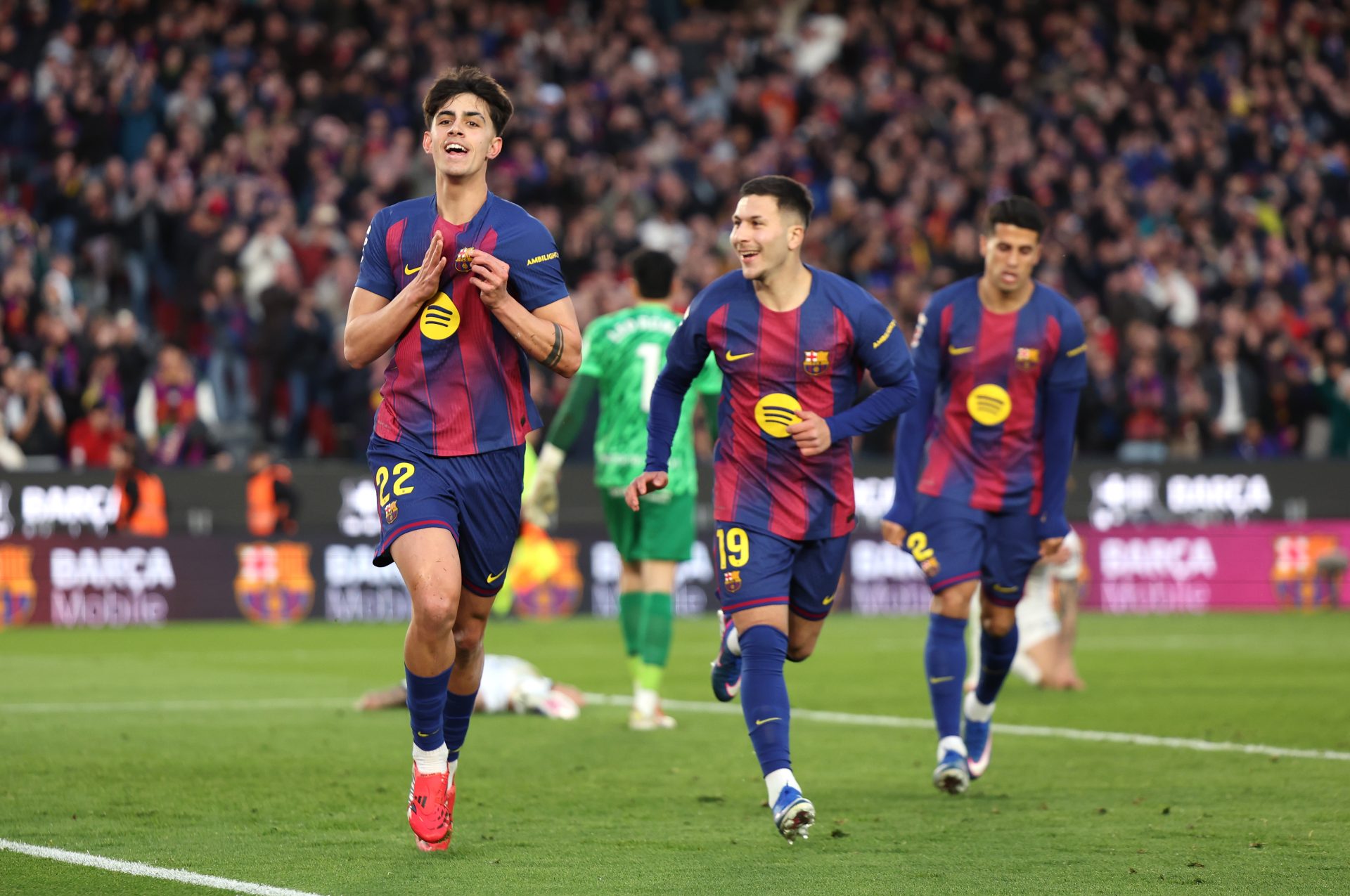 Tiga pemain FC Barcelona merayakan gol di lapangan rumput Stadion Olimpic Lluis Companys setelah pertandingan melawan RCD Mallorca. Pemain di tengah tersenyum lebar dan merangkul rekan setimnya, di saksikan oleh penonton di latar belakang.