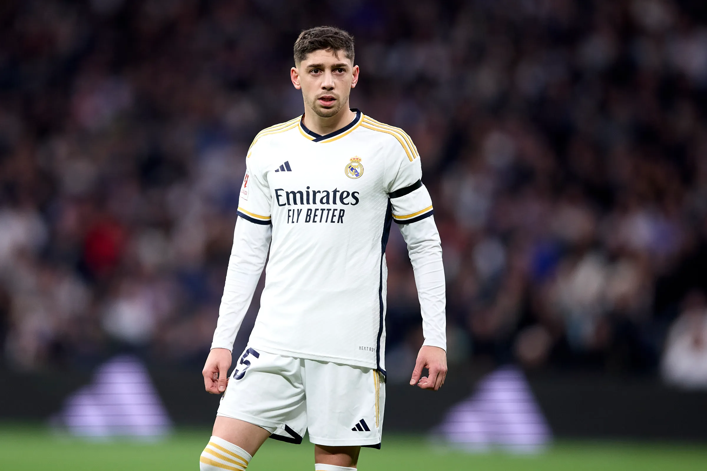 Federico Valverde dengan seragam kandang Real Madrid berwarna putih, berdiri di lapangan sepak bola dengan latar belakang tribun penonton yang buram.
