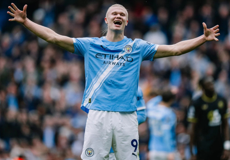 Seorang pemain Manchester City (kemungkinan Erling Haaland) merayakan gol dengan kedua tangan terangkat ke atas, di stadion, dalam pertandingan melawan Liverpool."