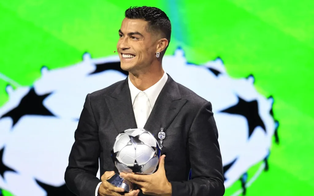 Cristiano Ronaldo memegang penghargaan pemain terbaik dengan latar belakang logo Liga Champions, mengenakan setelan jas hitam resmi.