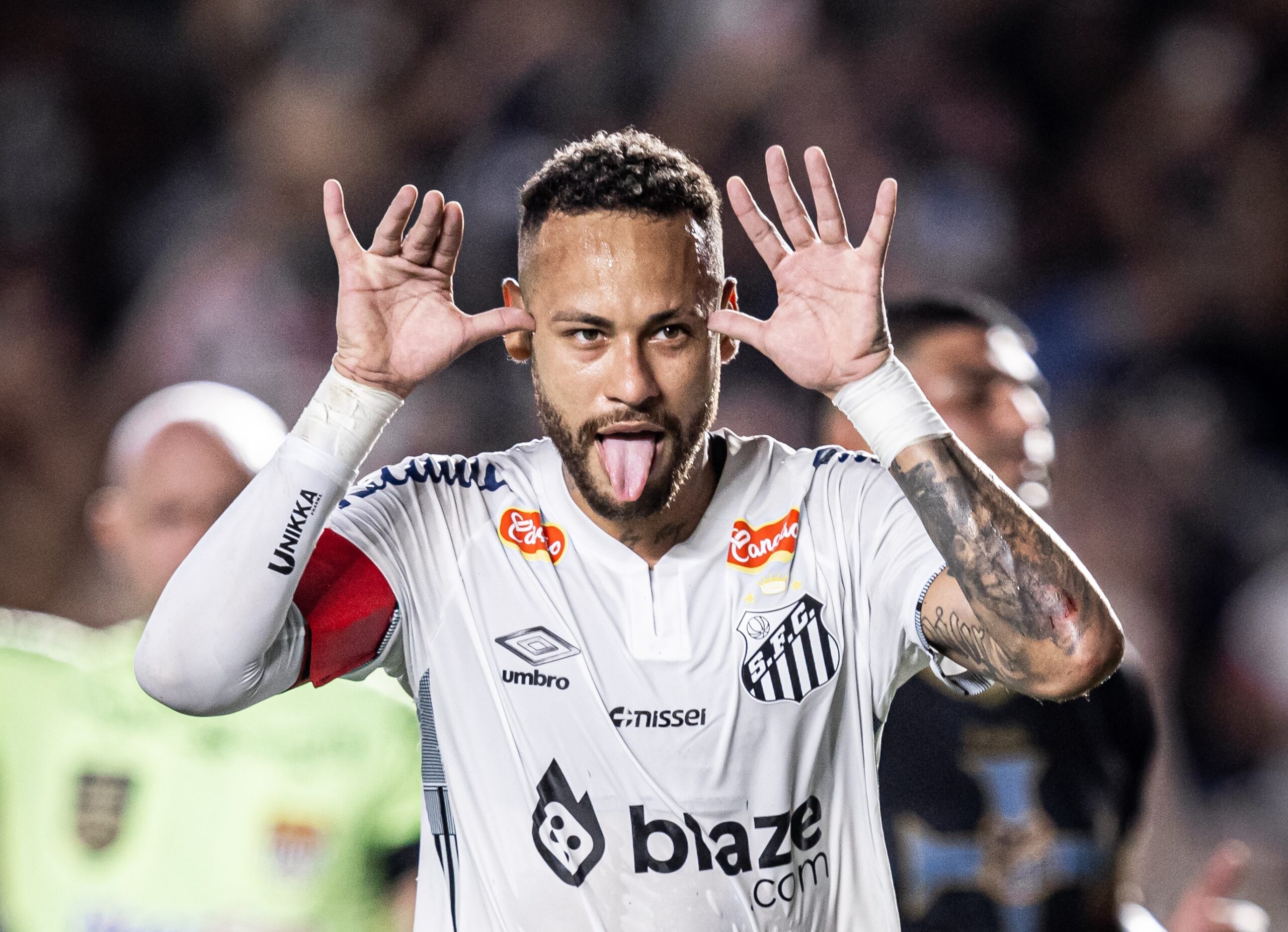 Pemain sepak bola Brasil, Neymar, menjulurkan lidah dan meletakkan tangan di samping telinganya dalam pose selebrasi saat mengenakan kostum putih Santos FC di lapangan sepak bola.