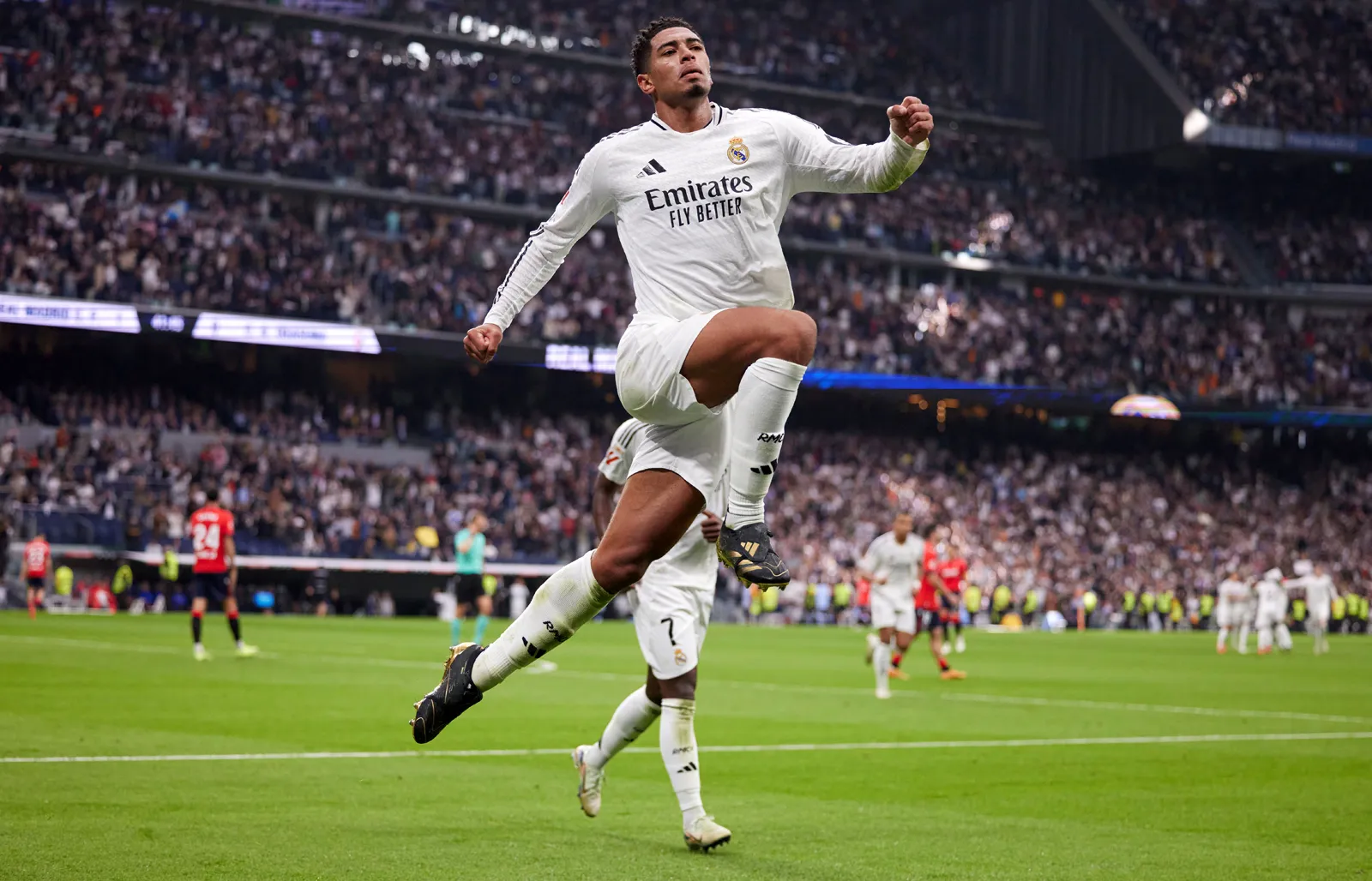 Jude Bellingham mengenakan jersei kandang Real Madrid berwarna putih, melompat di udara dengan satu lutut ditekuk dan kedua tangan terkepal di sebuah stadion yang penuh penonton, mengekspresikan kegembiraan setelah mencetak gol.