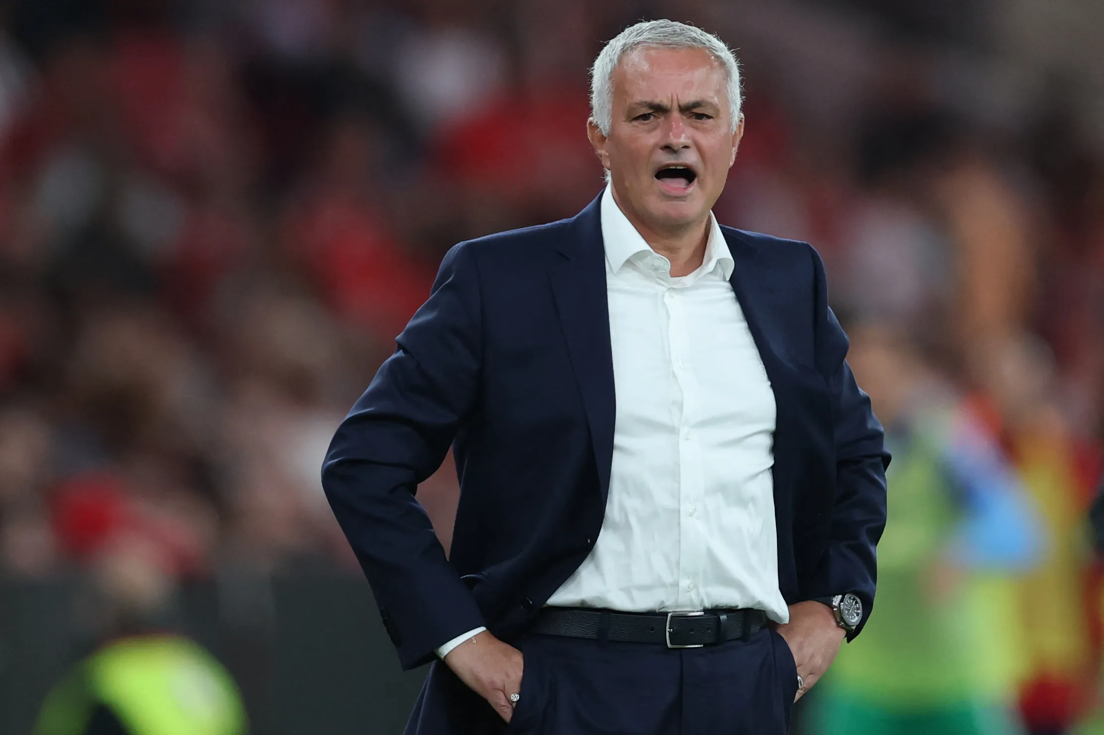 Jose Mourinho, pelatih kawakan dengan rambut putihnya yang khas, berdiri di pinggir lapangan mengenakan setelan jas.