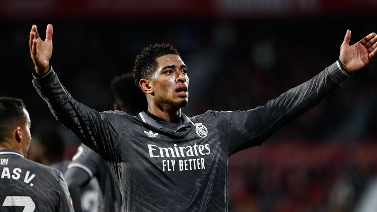 Potret setengah badan Jude Bellingham mengenakan jersei Real Madrid berwarna hitam, menengadahkan kepala dan mengangkat kedua tangan terbuka di sebuah stadion, seperti sedang merayakan sesuatu atau mengekspresikan emosi.