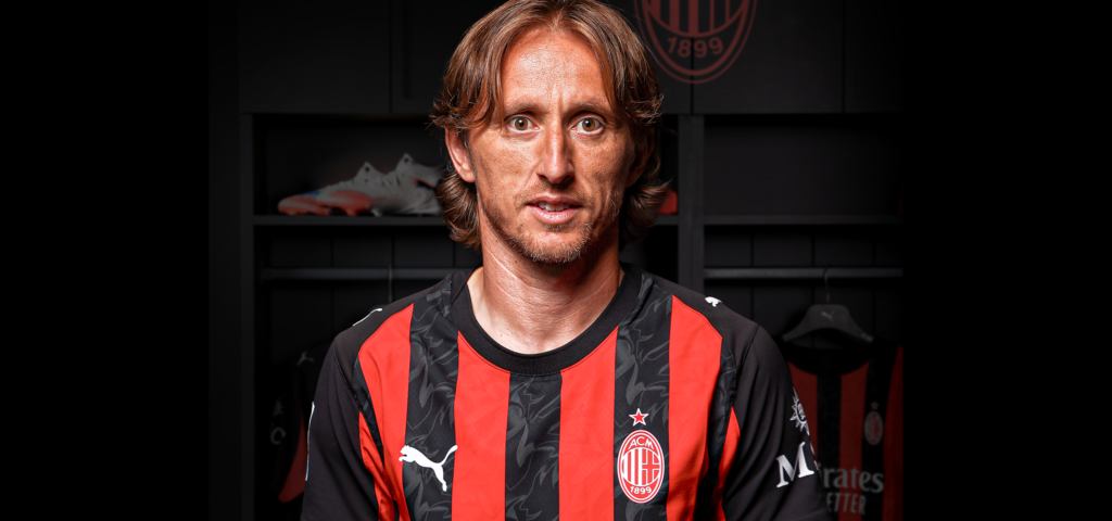Luka Modric mengenakan jersey AC Milan tersenyum menghadap kamera dengan latar belakang hitam.