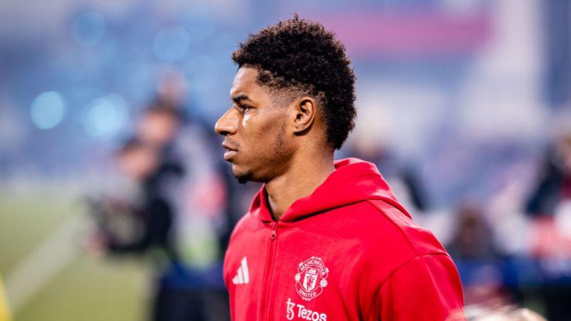 Marcus Rashford mengenakan jaket merah Manchester United tampak dari samping