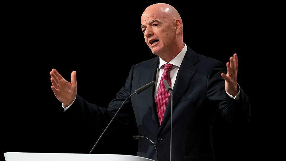 Presiden FIFA, Gianni Infantino, sedang berpidato di sebuah podium dengan kedua tangan terangkat ke samping, mengenakan setelan jas gelap dan dasi merah.