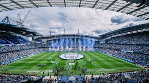 Suasana perayaan kemenangan Manchester City di Stadion Etihad dengan latar tribun biru dan bendera klub yang berkibar di pinggir lapangan.
