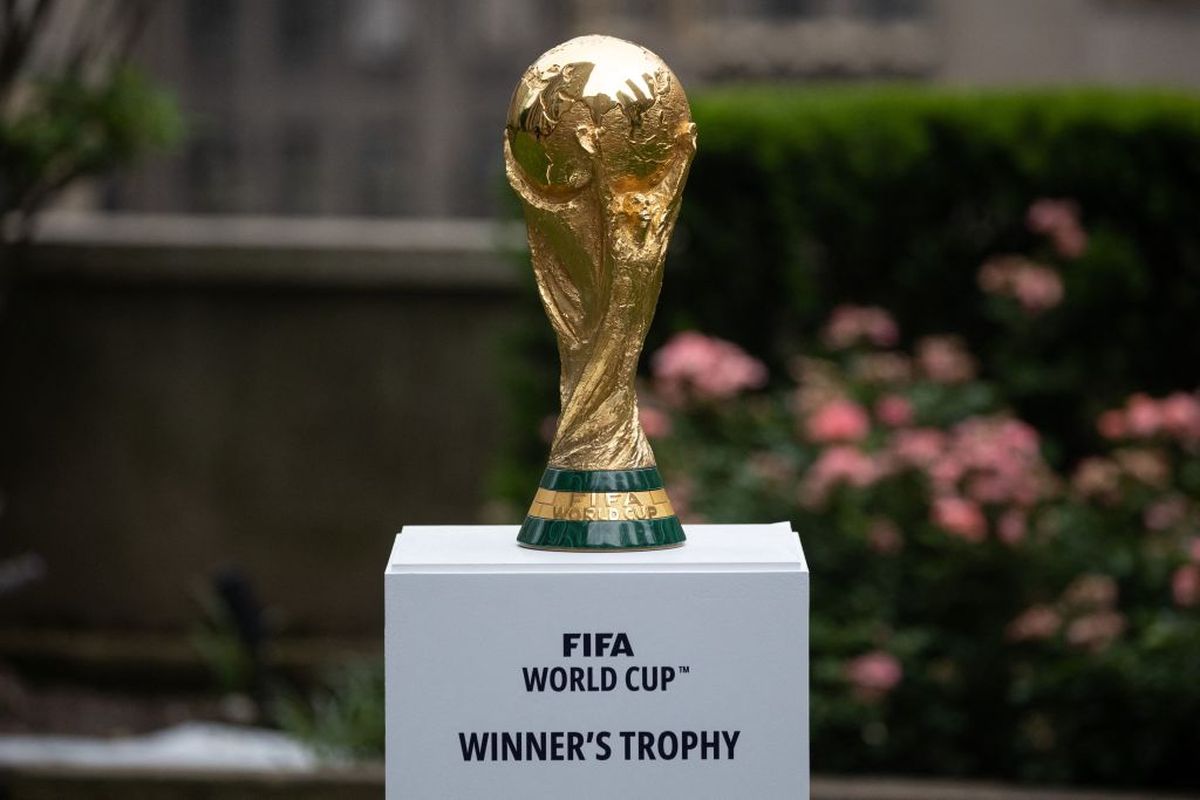 Foto trofi emas Piala Dunia FIFA yang diletakkan di atas podium putih dengan latar belakang taman dan bunga mawar