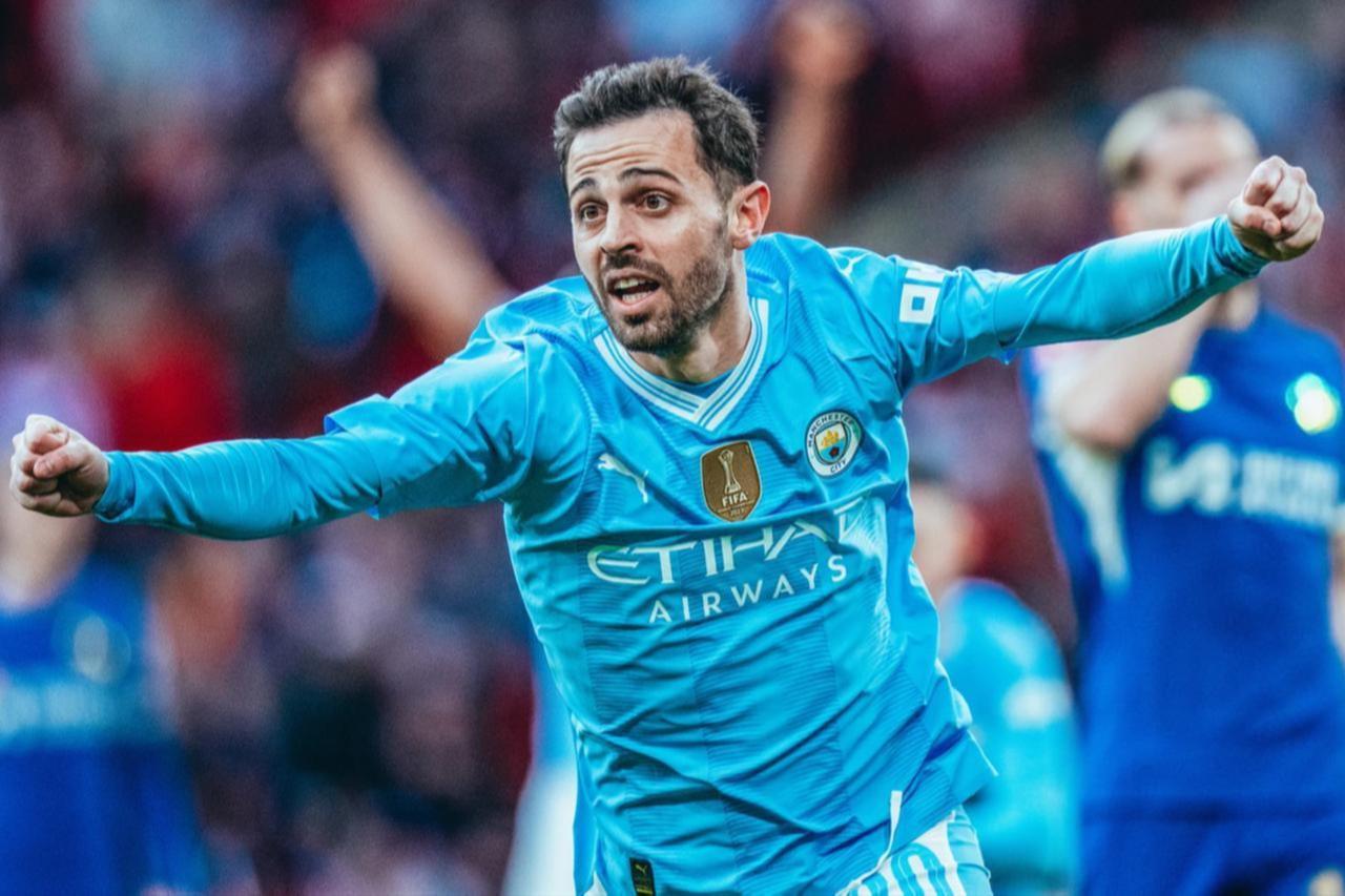 Bernardo Silva mengenakan jersi biru muda Manchester City sedang tersenyum lebar sambil merayakan gol di lapangan.
