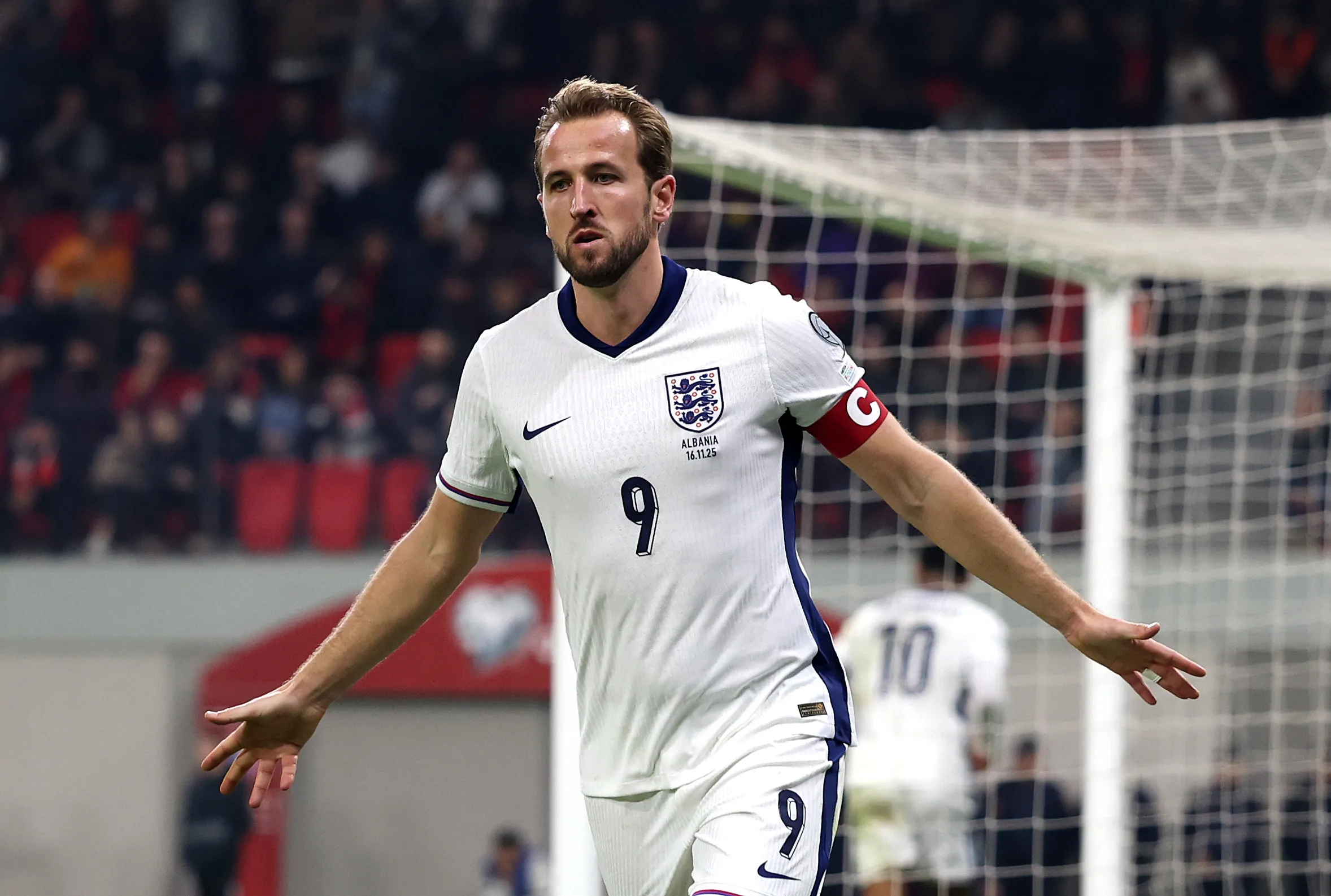 Striker Inggris Harry Kane merayakan gol dengan merentangkan kedua tangan memakai jersei putih bernomor 9 dan ban kapten di lengan.