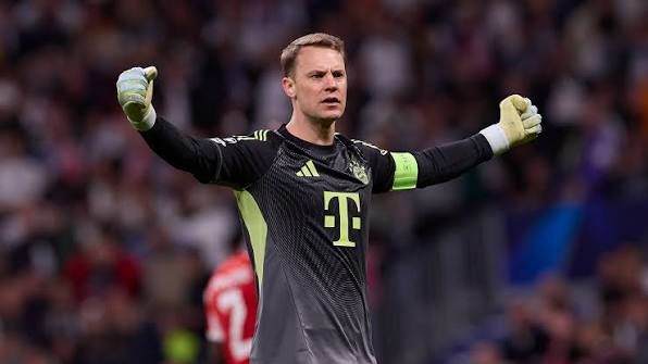 Manuel Neuer mengenakan seragam hitam Bayern Munchen dengan ban kapten, mengangkat kedua tangan merayakan kemenangan.