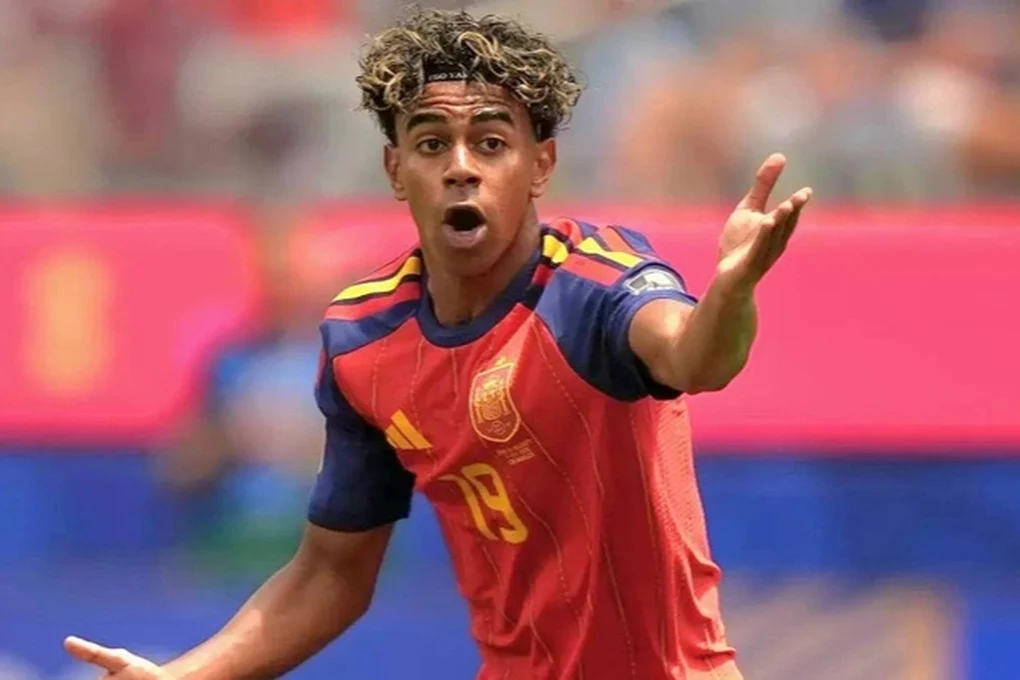 Lamine Yamal mengenakan jersey timnas Spanyol berwarna merah sedang merentangkan tangan dengan ekspresi wajah terkejut atau emosional di lapangan sepak bola.