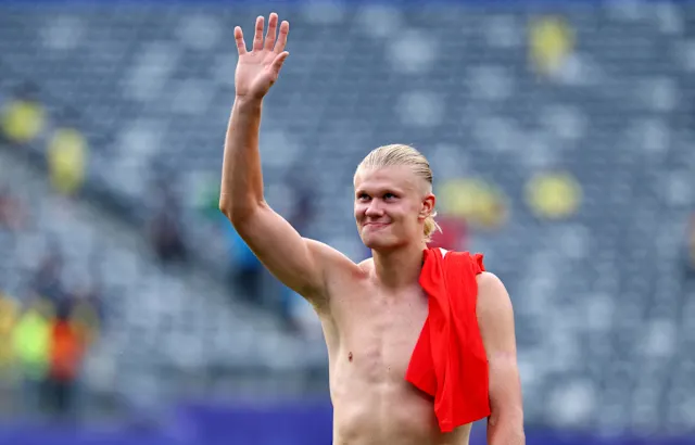 Erling Haaland tanpa mengenakan kaus sedang tersenyum dan melambaikan tangan kanan ke arah penonton di stadion, dengan jersi merah tersampir di pundak kirinya.