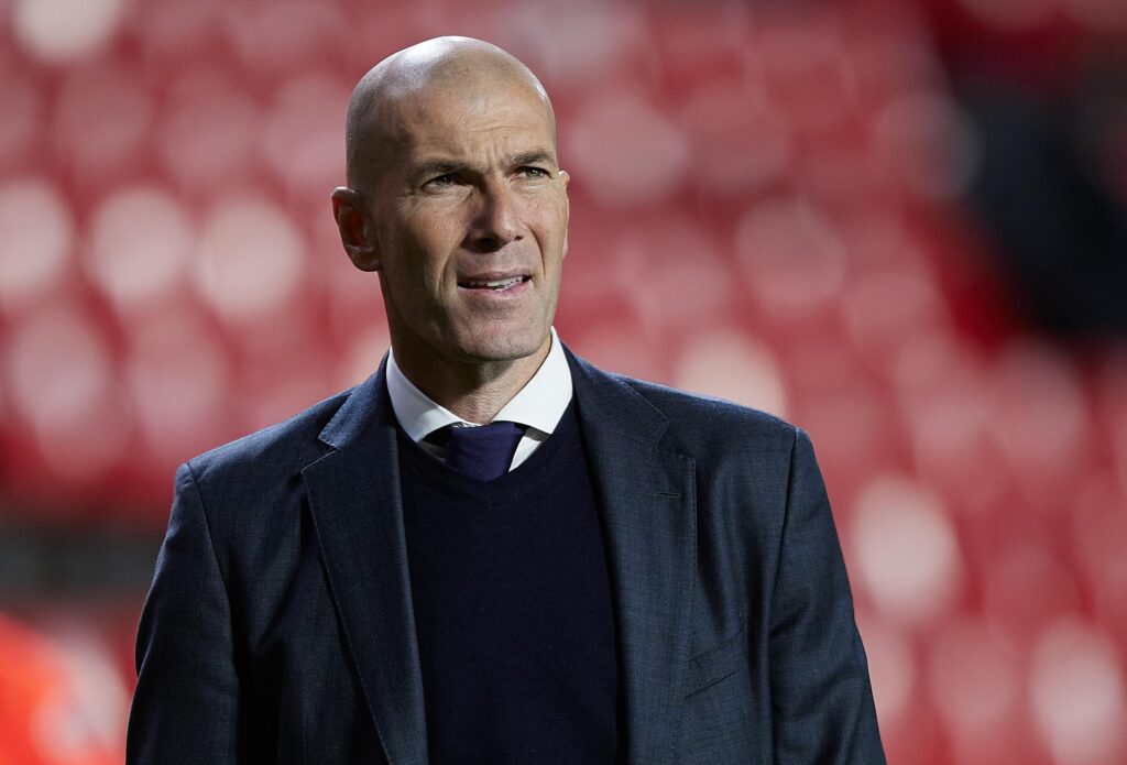 Potret Zinedine Zidane mengenakan jas hitam saat berada di pinggir lapangan.