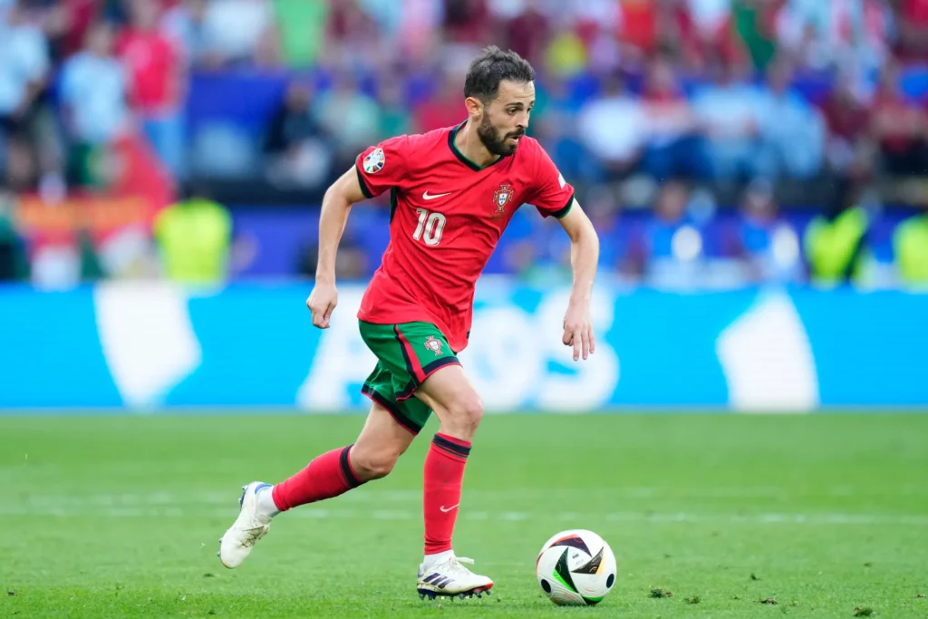 Foto yang menunjukkan seorang pemain sepak bola mengenakan jersei merah dan kaus kaki merah, serta celana pendek putih, sedang menggiring bola sepak di lapangan sepak bola.