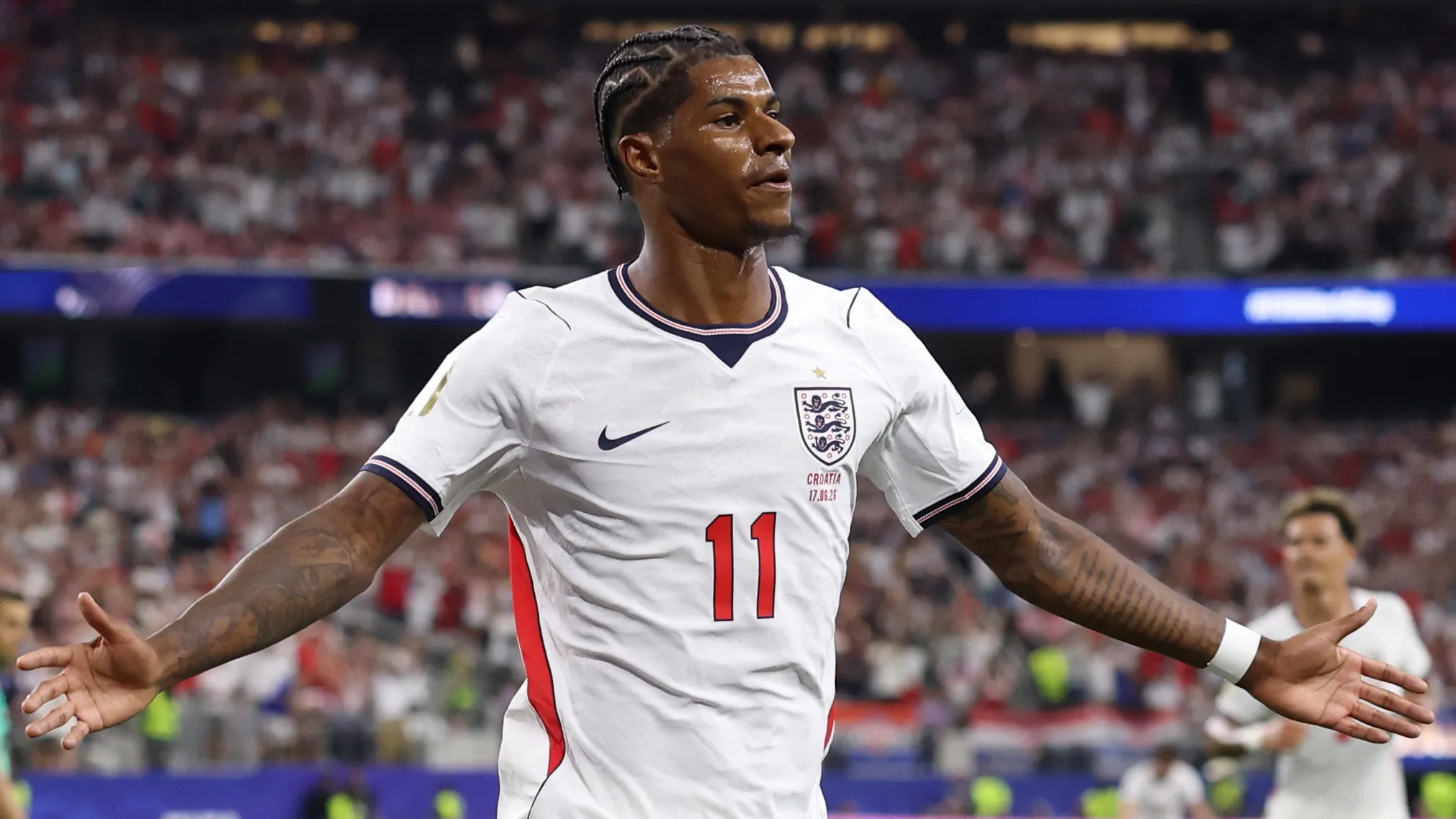 Pemain depan Timnas Inggris, Marcus Rashford, mengenakan jersei putih nomor 11 dan merentangkan tangannya di lapangan sepak bola saat melakukan selebrasi.