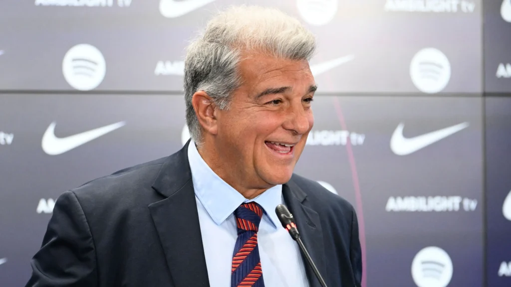 Joan Laporta mengenakan jas hitam dan dasi bercorak merah-biru sedang tersenyum dengan latar belakang logo Barcelona dan Nike.