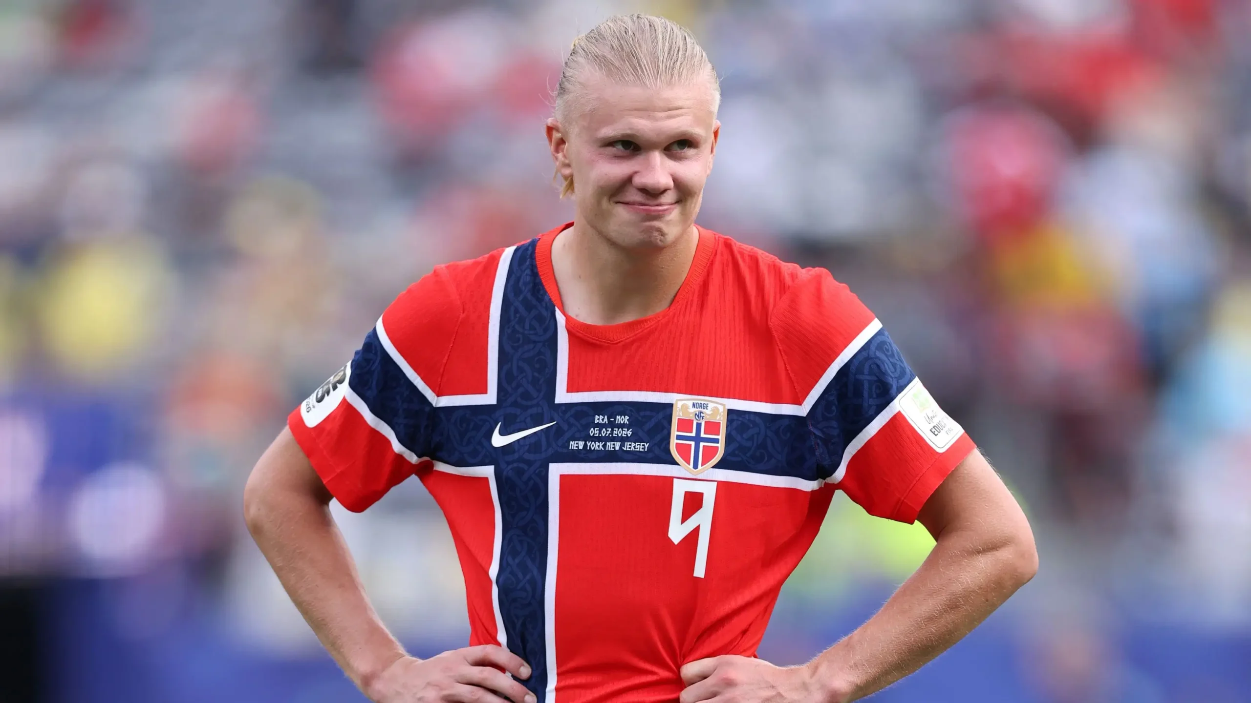 Erling Haaland mengenakan jersi merah tim nasional Norwegia, berdiri di lapangan dengan kedua tangan di pinggang dan menunjukkan ekspresi wajah lesu atau kecewa.