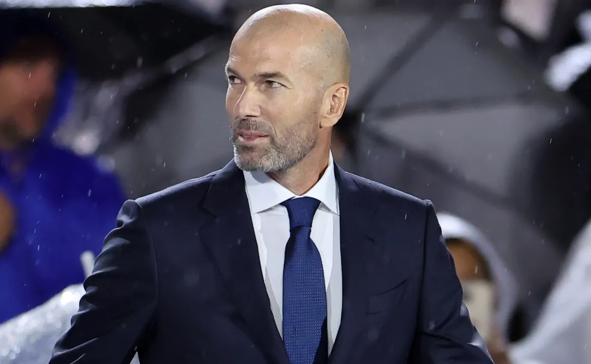 Zinedine Zidane mengenakan setelan jas biru tua dengan kemeja putih dan dasi senada saat melihat ke arah samping.