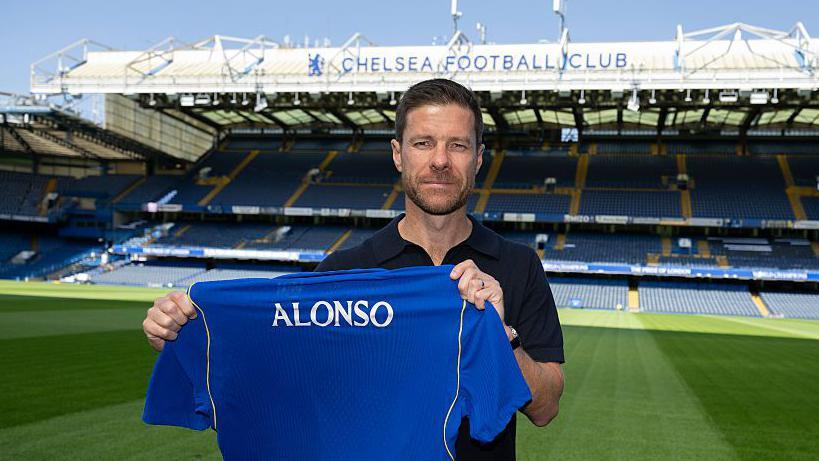 Foto Xabi Alonso memegang kaus Chelsea FC dengan nama 'ALONSO' di pinggir lapangan Stamford Bridge
