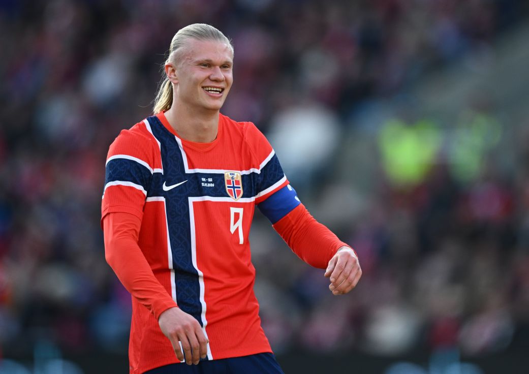 Erling Haaland mengenakan jersi merah lengan panjang timnas Norwegia, sedang berjalan di lapangan hijau saat pertandingan dengan ekspresi wajah serius dan fokus.