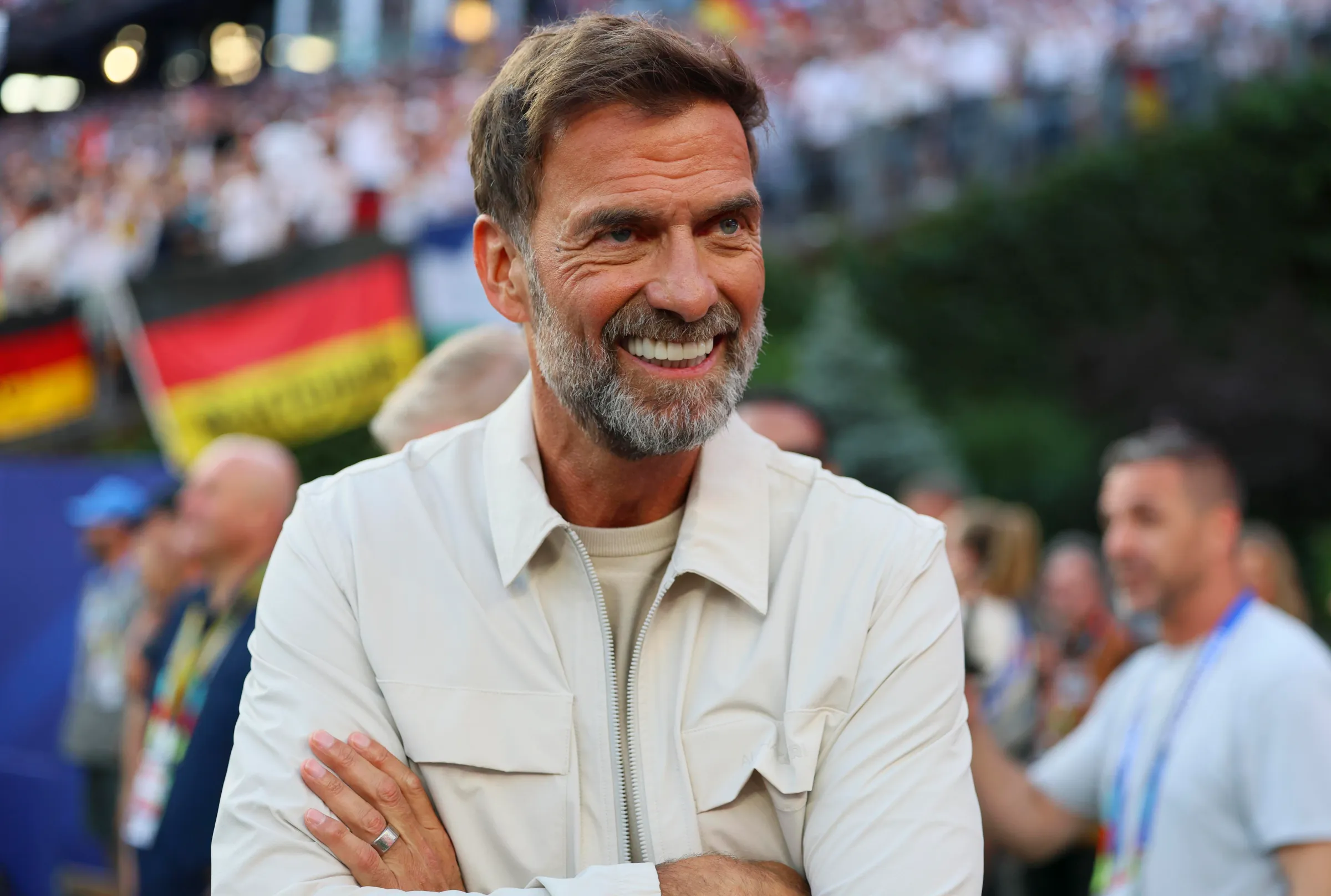 Jurgen Klopp tersenyum di tribun stadion dengan latar belakang suporter membawa bendera Jerman