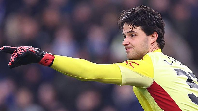 Senne Lammens menunjuk ke arah lapangan saat bertanding mengenakan seragam kiper kuning.