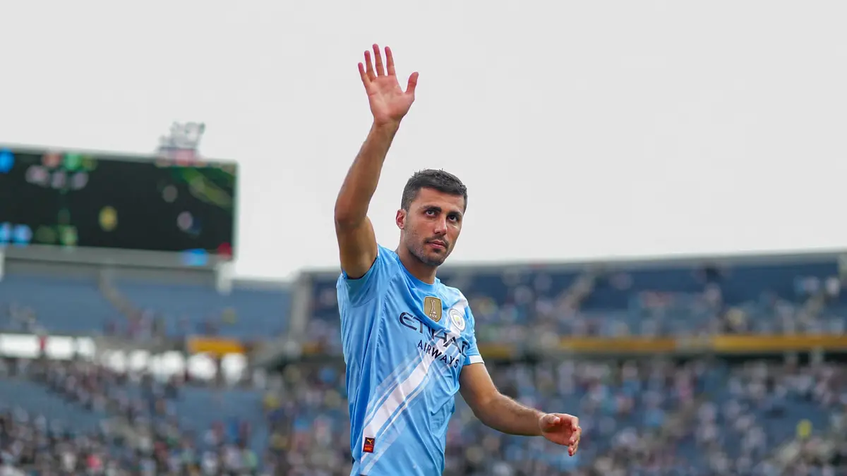 Rodri mengenakan seragam biru muda Manchester City, melambaikan tangan ke arah penonton di stadion.