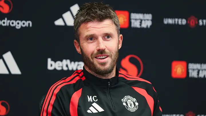 Michael Carrick, pelatih Manchester United, tersenyum dalam balutan jaket latihan hitam berlogo adidas dan Manchester United.